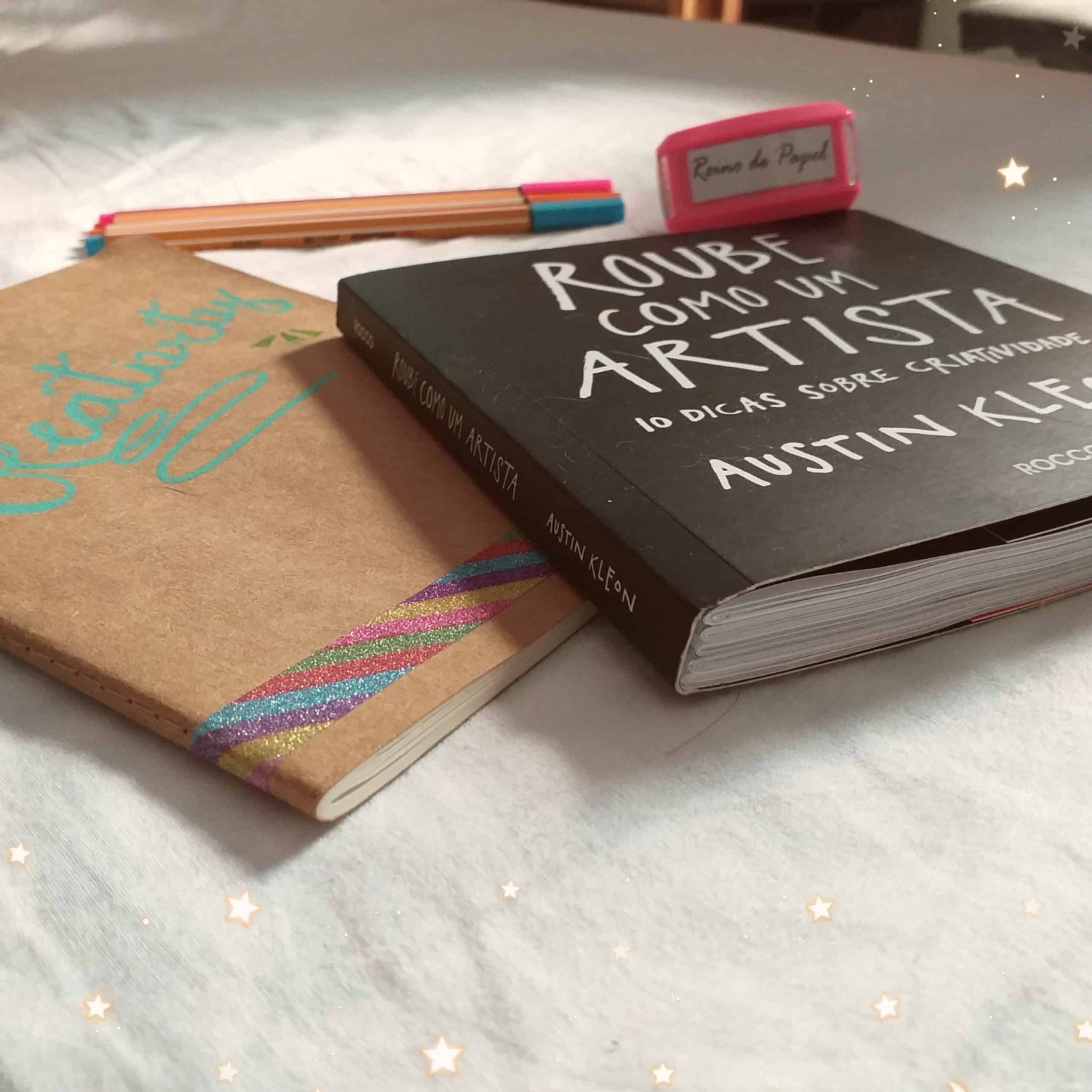 Vista periférica do livro Roube como um artista, ao lado de um caderno com capa marrom canetas coloridas e um carimbo escrito Reino de Papel